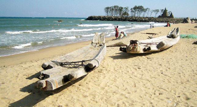 Two wooden canoes resting on a sandy beach near turquoise ocean waves, with people walking in the distance under a clear sky